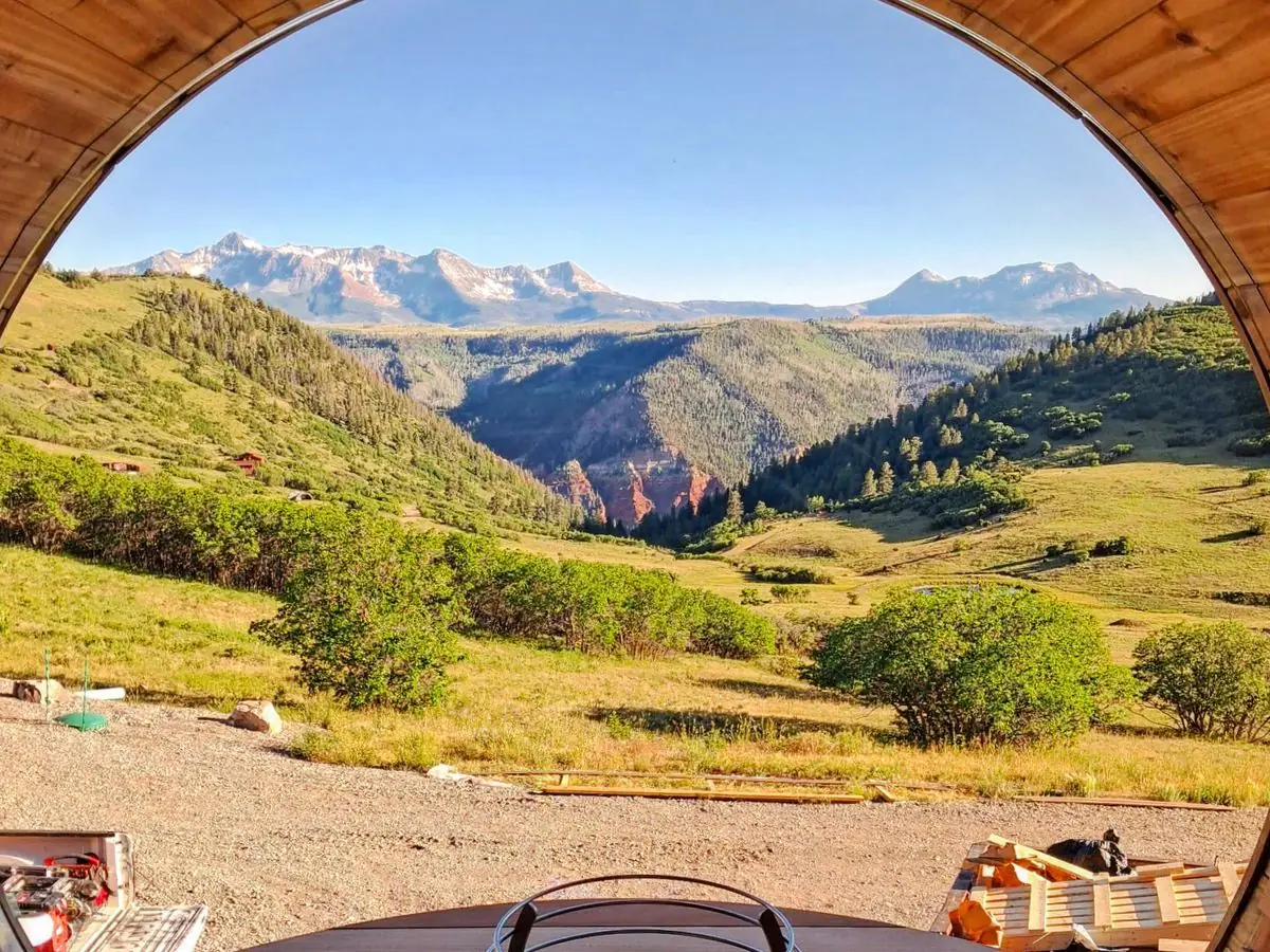 Inside a cedar barrel sauna with a HUUM heater, looking out a panoramic window at a mountain valley